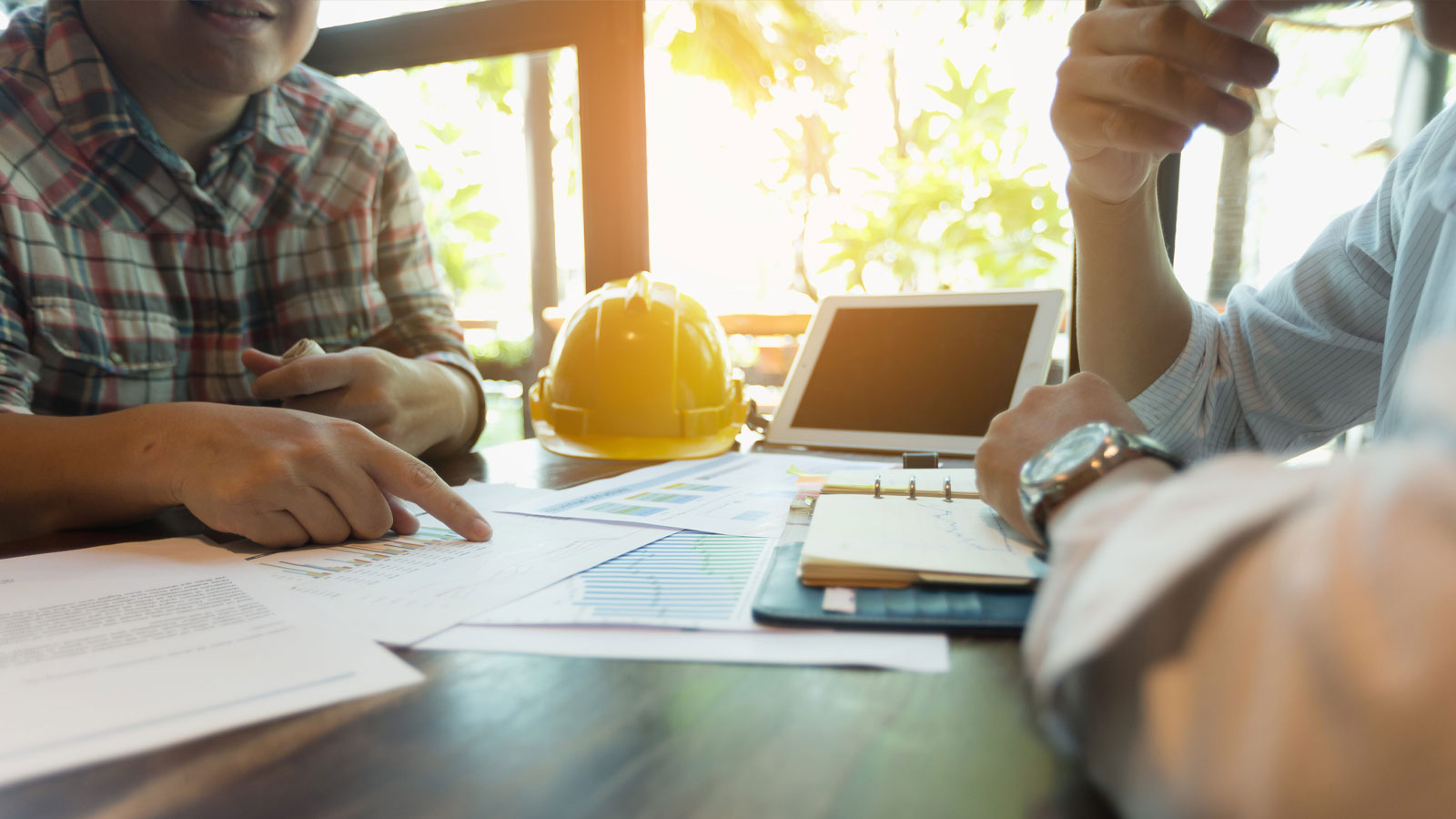 Top view of an architect and an engineer wearing safety helmets looking at blueprints in the middle of a construction site, surrounded by construction workers and building materials.